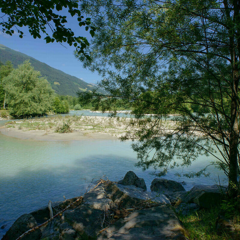 Das türkis-blaue Wasser der unteren Isel in Oberlienz. In der Mitte ist eine kleine Sandbank. Am Ufer wachsen viele grüne Bäume.