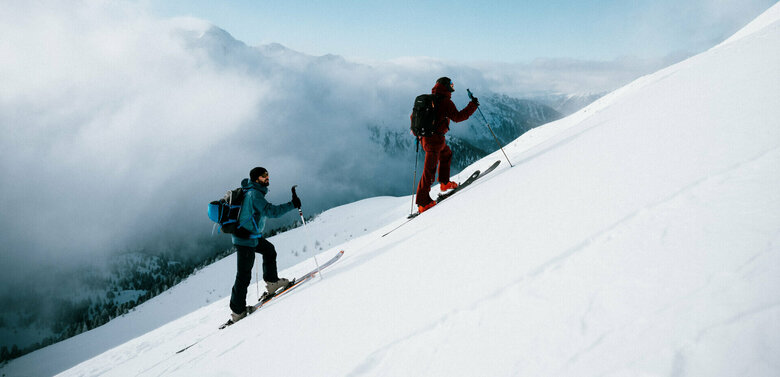 Zwei Skitourensportler erklimmen im Gleichschritt auf ihren Tourenskiern einen mittelsteilen Berghang an einem Schönwettermorgen. Dahinter verdeckt zäher Hochnebel einen Teil der Berge.