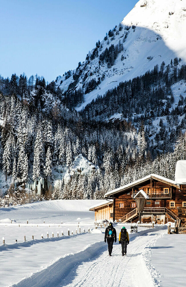 Eiskletterpark Osttirol Zwei Personen spazieren auf einem Winterwanderweg an einer kleinen Ansammlung von kleinen Hütten vorbei. Dahinter der bewaldete Talschluss in einer winterlichen Umgebung.