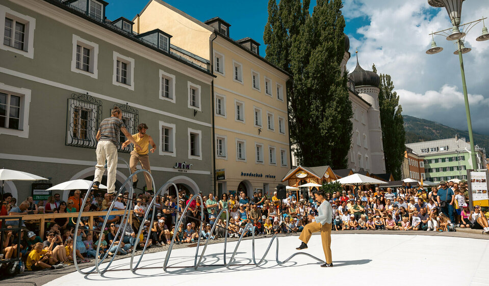 Drei Artisten turnen auf, in bzw. vor einer großen Spirale aus Eisen am Lienzer Hauptplatz im Rahmen von Olala 2025. Sie werden vom begeisterten Publikum rundherum bestaunt.