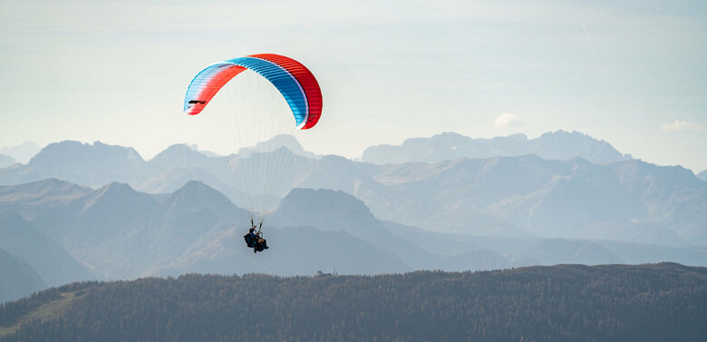 Ein Gleitschirm mit blau-roter Färbung fliegt hoch über Osttirol, im Hintergrund sind die imposanten Berge zu sehen.