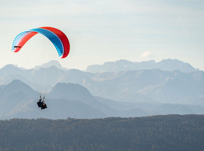 Ein Gleitschirm mit blau-roter Färbung fliegt hoch über Osttirol, im Hintergrund sind die imposanten Berge zu sehen.