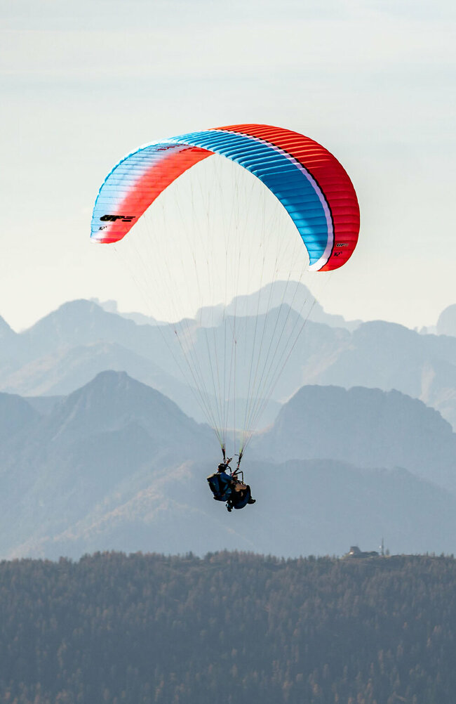 Ein Gleitschirm mit blau-roter Färbung fliegt hoch über Osttirol, im Hintergrund sind die imposanten Berge zu sehen.
