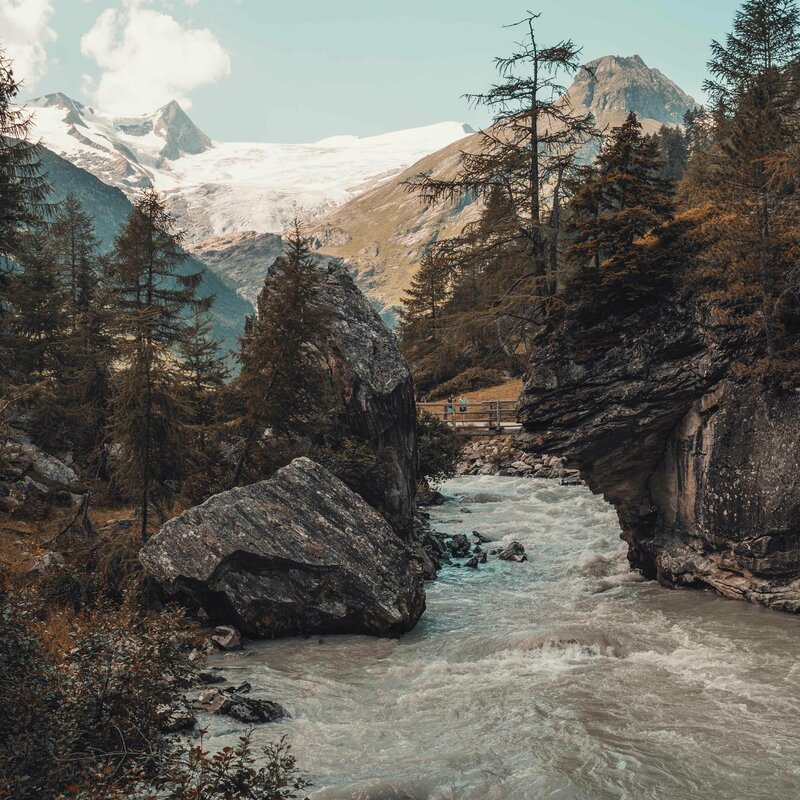Aufnahme von einem schnell fließenden Fluss, umgeben von großen Felsen und einigen Nadelbäumen. Im Hintergrund ist eine kleine Holzbrücke mit 2 Personen und es erheben sich hohe, noch schneebedeckte Berge.
