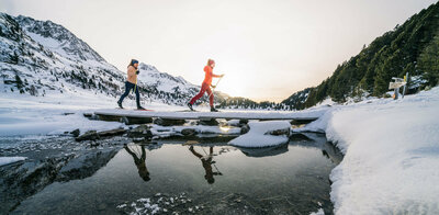 zwei Langläufer:innen überqueren einen Bach beim Langlaufen im Defereggental