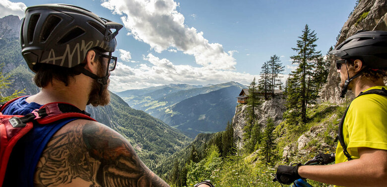 Zwei Männder mit Helmen halten ihre Mountainbikes und blicken auf die Dolomitenhütte auf dem Felsvorsprung.