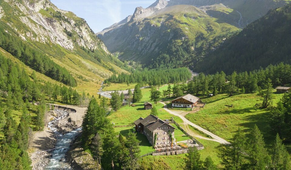 Blick auf das Kalser Tauernhaus auf der zweiten Etappe der Glocknerkrone in Osttirol. Grüne Almen, der Dorferbach und die Glocknergruppe im Hintergrund.