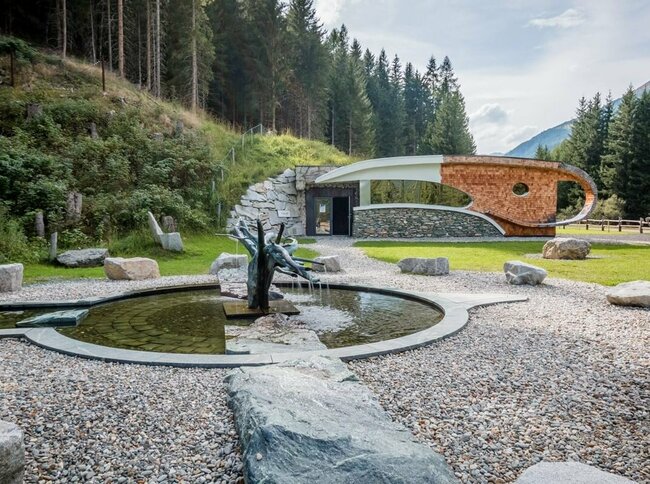 Der Außenbereich der Deferegger Heilwasserwelt in St. Jakob in Defereggen. Der Brunnen gefüllt mit Heilwasser ist der Mittelpunkt des Bildes, dieser ist umgeben von Kieselsteinen. Im Hintergrund ist die sogenannte "Serona-Höhle" zu sehen, welche von grünem Gras und einem dichten Wald umgeben ist.