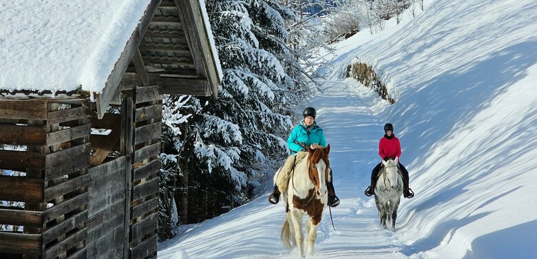 Zwei Frauen reiten mit einem braun-weißen und einem grauen Pferd auf einem schneebedeckten Weg in Prägraten. Links steht eine schneebedeckte, hölzerne Hütte.