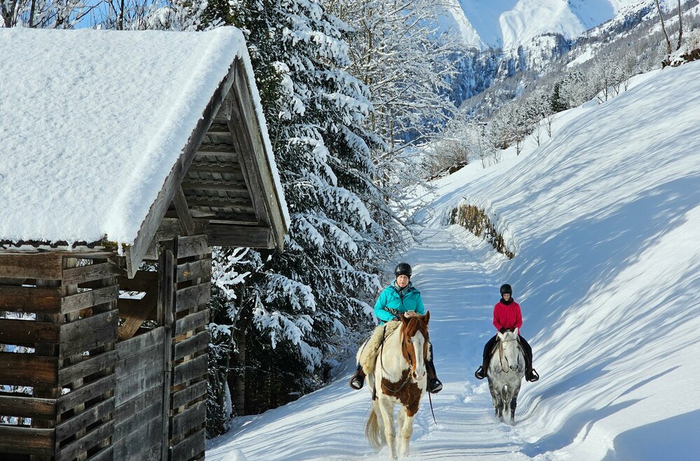Zwei Frauen reiten mit einem braun-weißen und einem grauen Pferd auf einem schneebedeckten Weg in Prägraten. Links steht eine schneebedeckte, hölzerne Hütte.