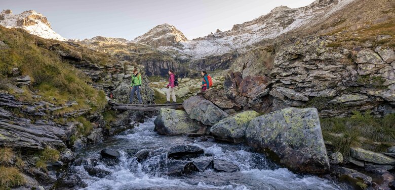 Drei Frauen überqueren auf einem schmalen Steg einen klaren Bach im Hochgebirge, umgeben von mächtigen Gipfeln