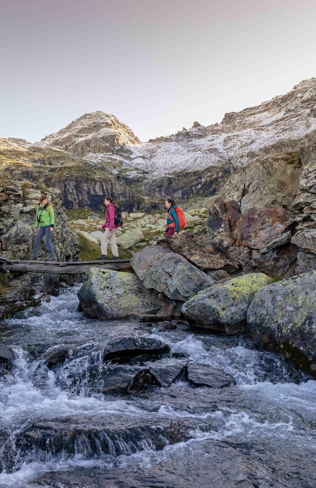Herbstwanderung mit Ranger Drei Frauen überqueren auf einem schmalen Steg einen klaren Bach im Hochgebirge, umgeben von mächtigen Gipfeln