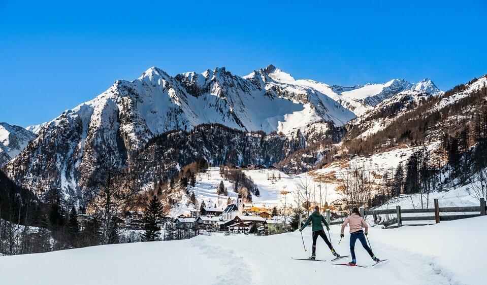 Zwei Langläuferinnen von hinten skaten in Richtung Dorf in Prägraten am Großvenediger. Im Hintergrund erheben sich schneebedeckte hohe Berge in den blauen Himmel.
