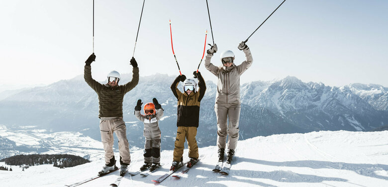 Familie mit zwei Kindern auf Skiern im Skigebiet Zettersfeld in Osttirol. Gemeinsam strecken sie jubelnd die Arme und Skistöcke in die Höhe, vor dem Panorama der verschneiten Lienzer Dolomiten.