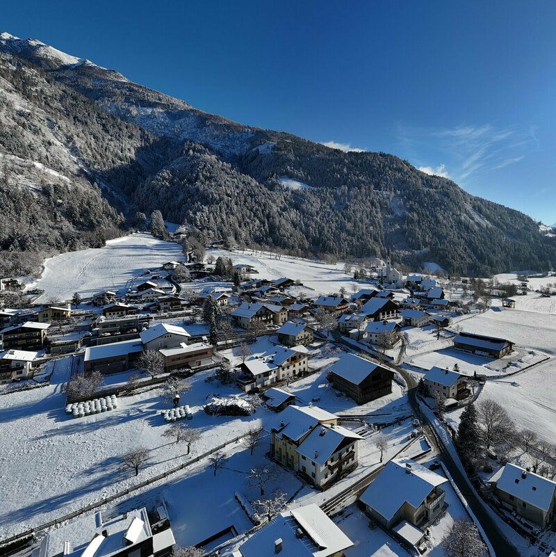 This panoramic photo shows a winter landscape of the village of Ainet in Osttirol. The roofs of the numerous houses, along with the village church and the streets, are largely covered in snow. The village is bordered by white, snow-covered fields and meadows.