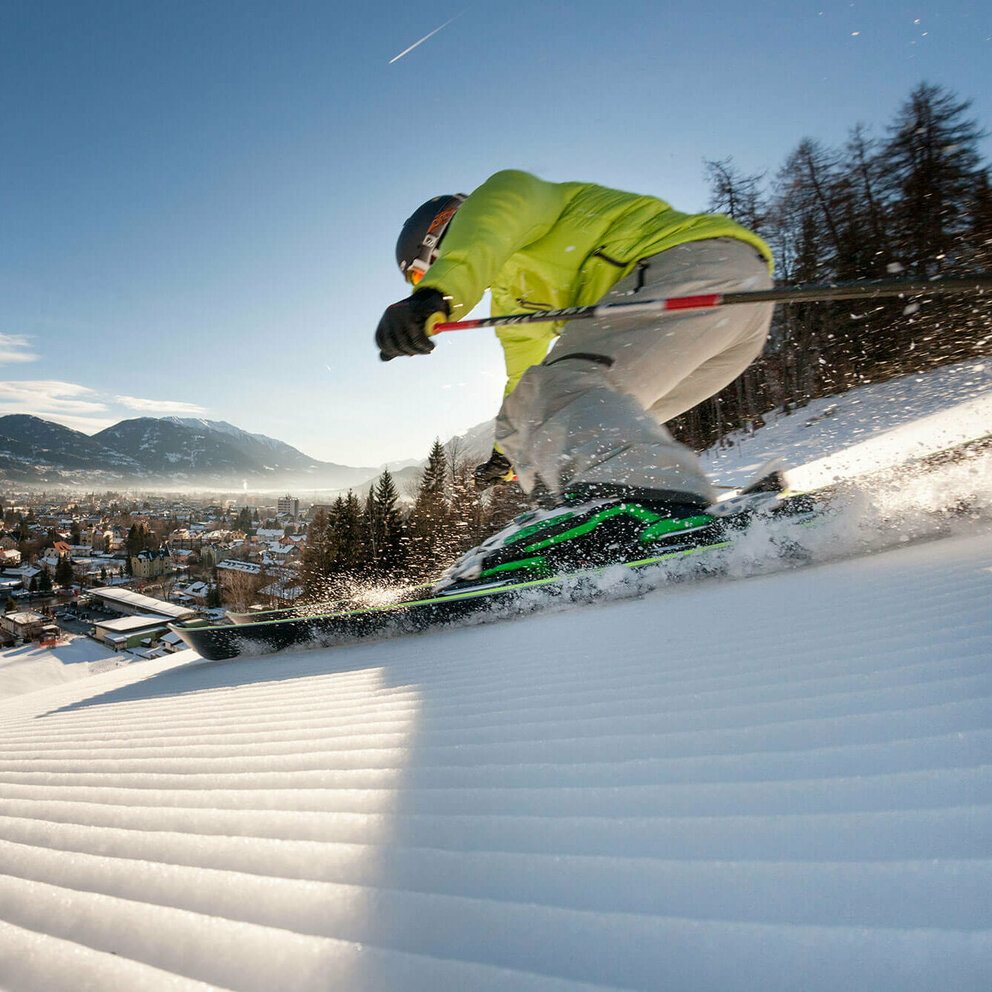 Ein Skifahrer mit gelber Jacke und heller Hose carved am Hochstein mit den Ski. Aufgenommen von unten. Blick auf das Dorf.