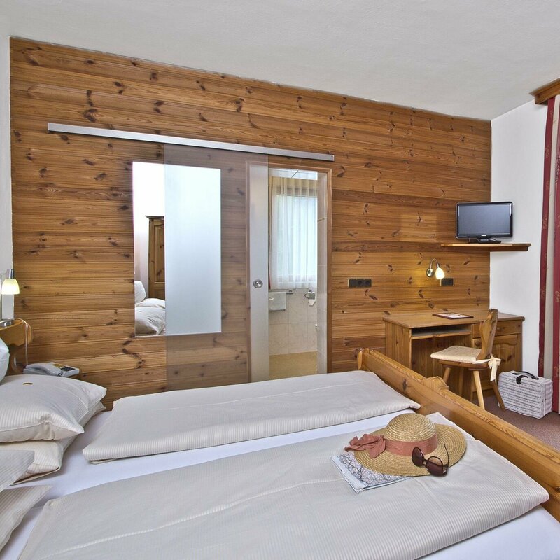 Ein Zimmer im Hotel Laserz mit Bett, Tisch, TV, Badezimmer und Balkon. Auf dem Bett liegt ein Hut, eine Landkarte und eine Sonnenbrille.