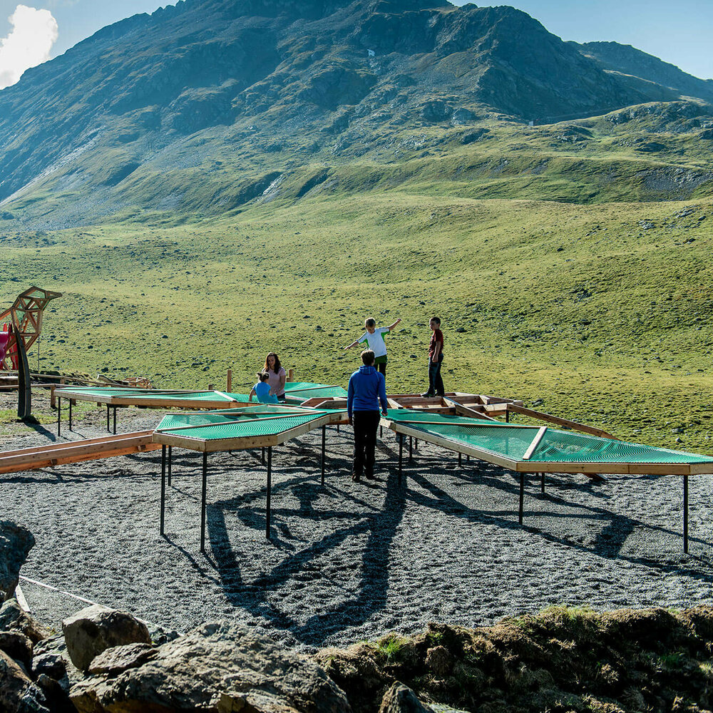 Spielende Kinder beim Wassermythos Ochsenlacke im Skizentrum St. Jakob i. D.. Die Kinder toben auf einem der vielen Spielgeräte während die Sonne die Umgebung und die dahinterliegende Bergwelt in einem warmen Licht erstrahlen lässt.