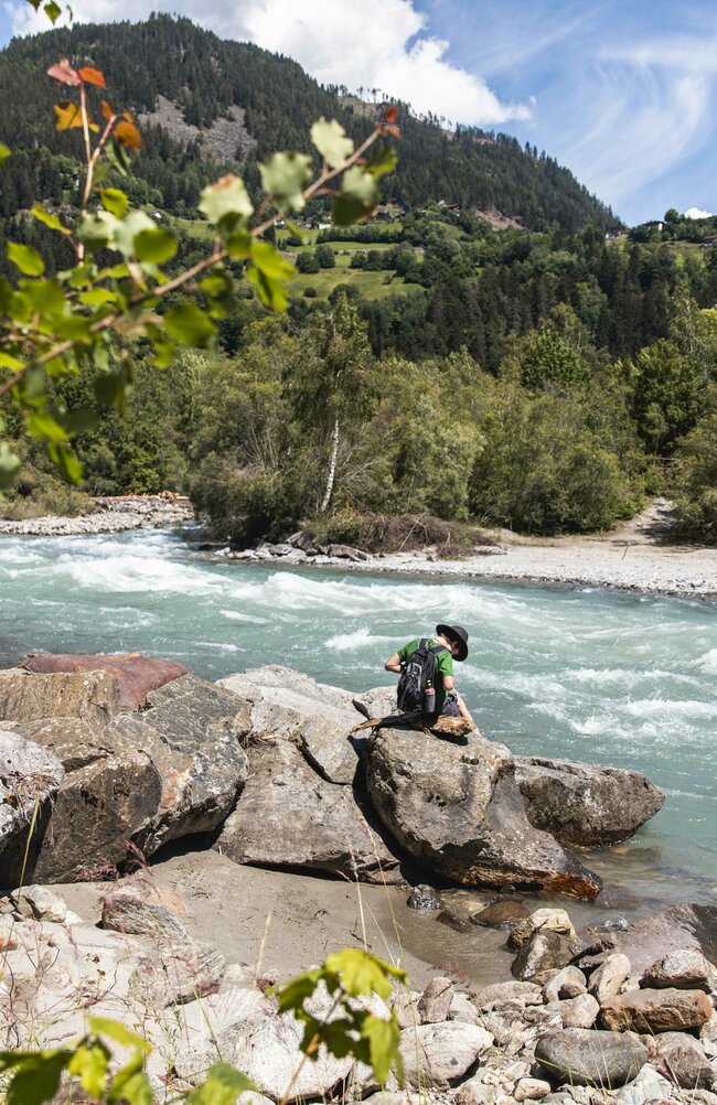 Iseltrail verläuft direkt an der Isel vorbei mit Wäldern am Ufer. Ein Mann mit grünem Shirt und Rucksack sitzt auf einem Felsen direkt am Wasser.