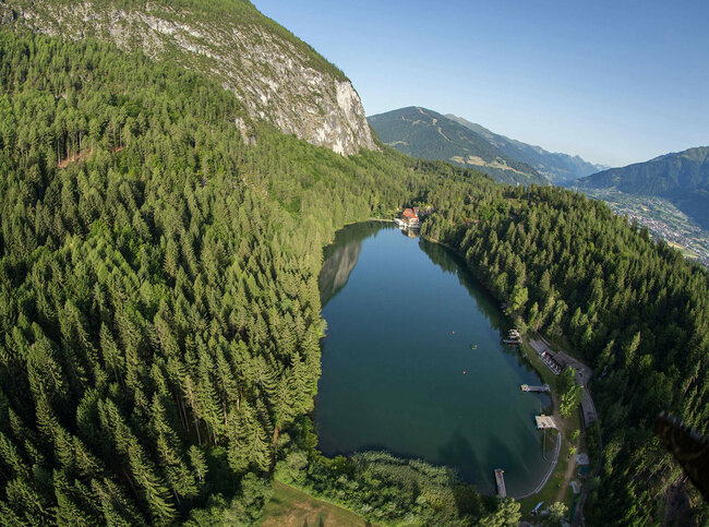 Luftaufnahme vom sanft in den Wald eingebetteten Tristacher See bei herrlichem Wetter, mit saftig grünen Bäumen drum herum.