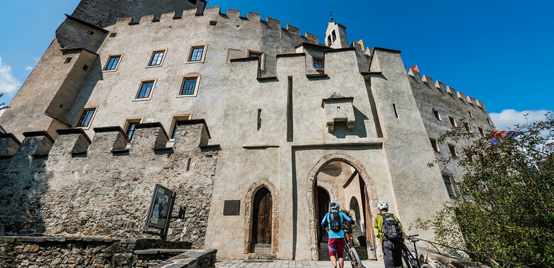 Zwei Biker schieben ihre Räder durch einen steinernen Bogen am Eingang des Schloss Bruck in Lienz.
