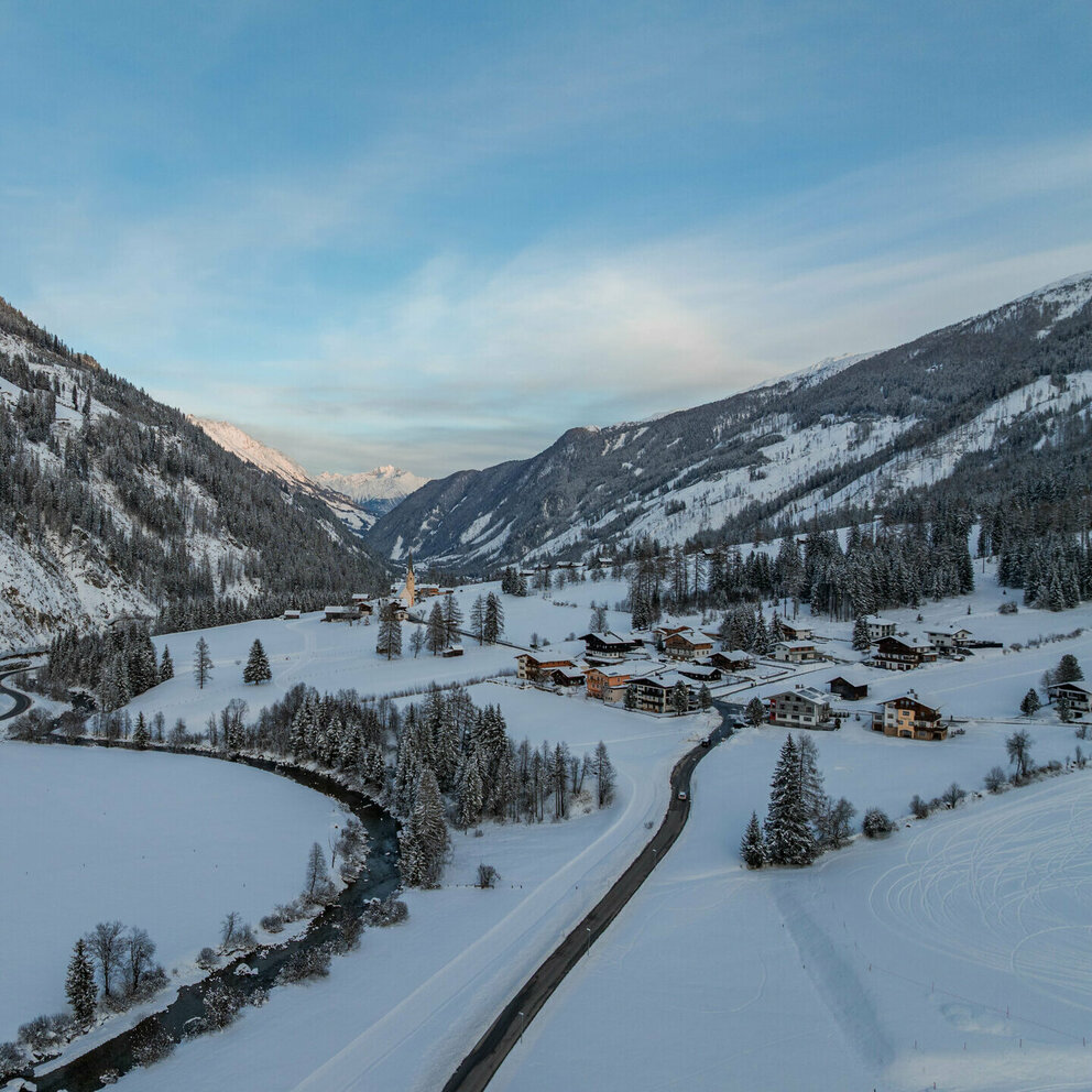 Luftaufnahme eines verschneiten Alpentals mit Fluss, Straßen, einem kleinen Dorf und bewaldeten Berghängen im Winterlicht.