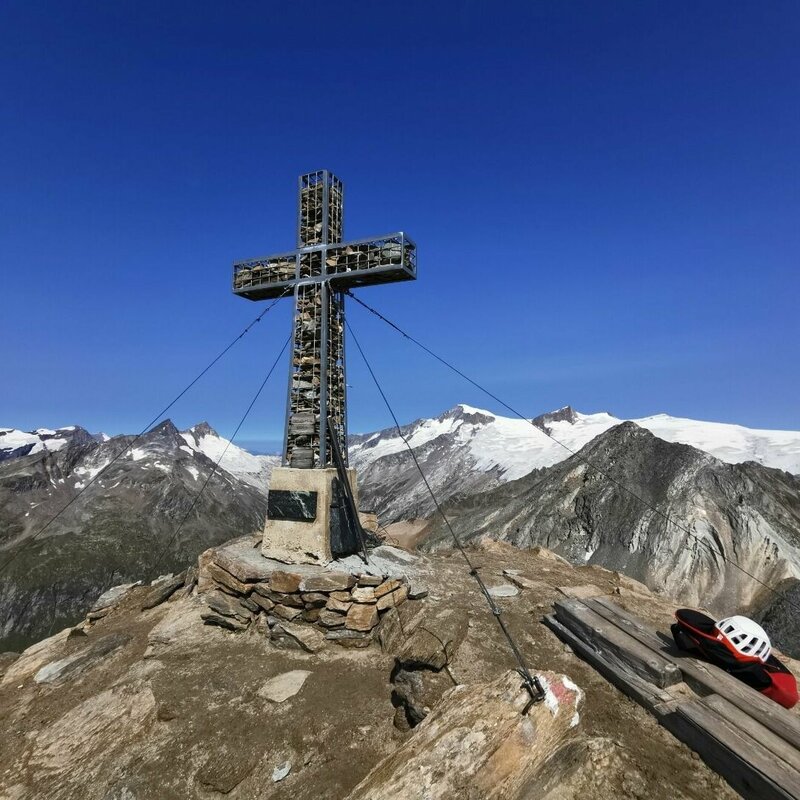 Das eindrucksvolle Gipfelkreuz der Kreuzspitze. Ein mit Steinen gefüllter Rahmen steht mächtig im blauen Himmel.