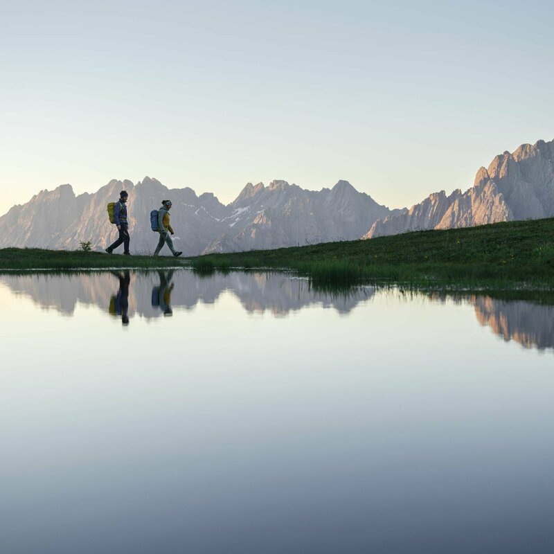 Ein Paar wandert um einen See bei einer Sonnenaufgangstour, waehrend sich die Berge im See spiegeln.