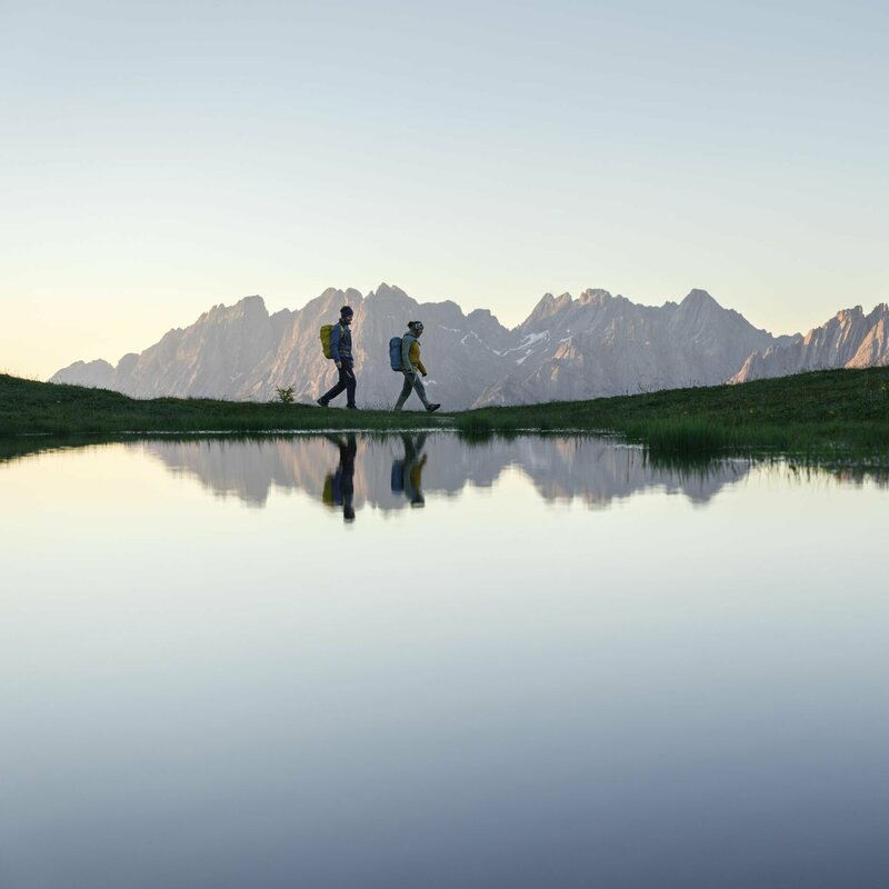 Ein Paar wandert um einen See bei einer Sonnenaufgangstour, waehrend sich die Berge im See spiegeln.