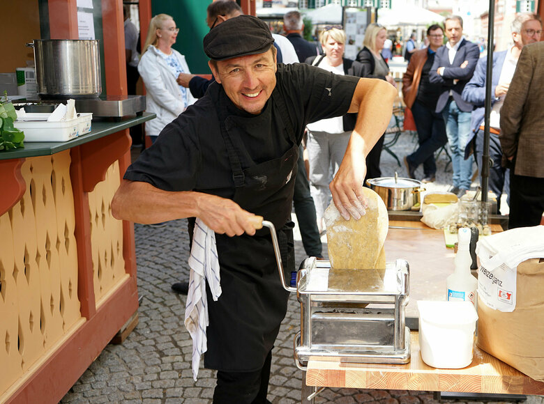 Koch Ernst Muhr dreht auf seinem Stand den Nudelteig durch die Nudelmaschine. Einige Menschen sehen ihm dabei zu.