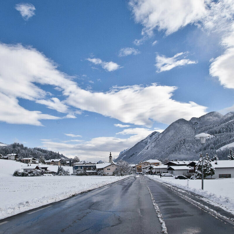 Straße, die in den Ort Abfaltersbach führt, das schon im Hintergrund zu erkennen ist. Neben der Fahrbahn liegt Schnee.