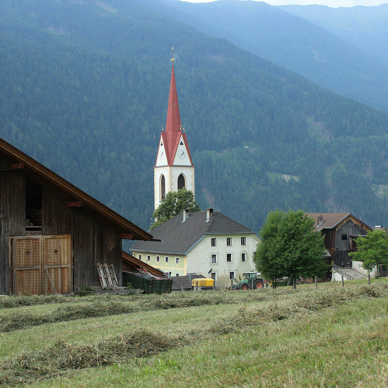 Ein Bauernhof in Oberlienz mit dem Kirchtum der Gemeinde im Hintergrund.