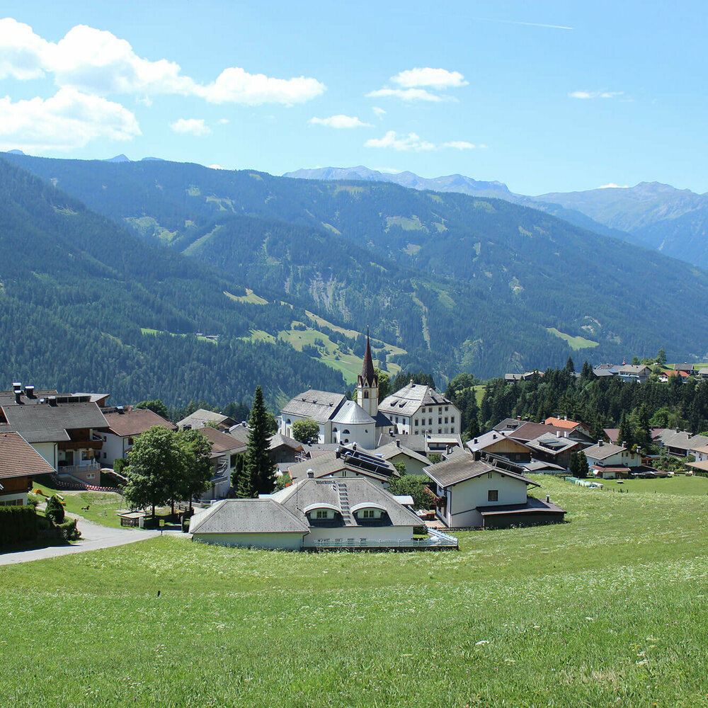 Blick auf Anras mit einem grünem Feld im Vordergrund und bewaldeten Berghängen im Hintergrund.