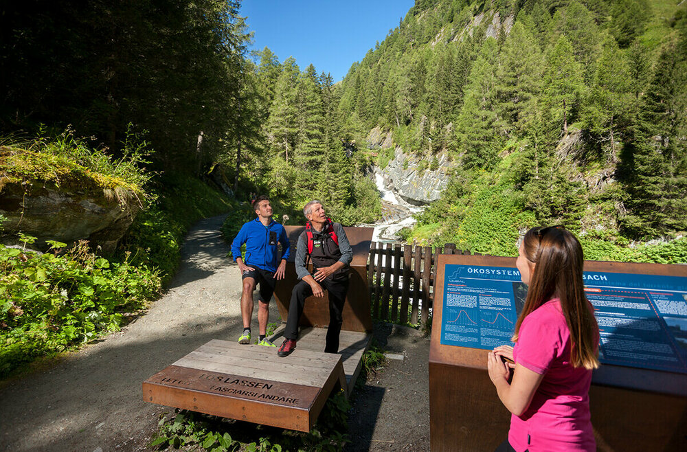 An einem Sommertag im Nationalpark Hohe Tauern betrachten drei Personen eine Informationstafel, während im Hintergrund die Isel als Gletscherfluss durch die Landschaft fließt