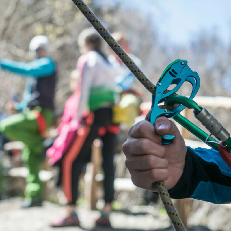 Nahaufnahme von einem Kletter-Sicherungsgerät, einem Seil und bunten Karabinern. Im Hintergrund stehen mehrere Personen.
