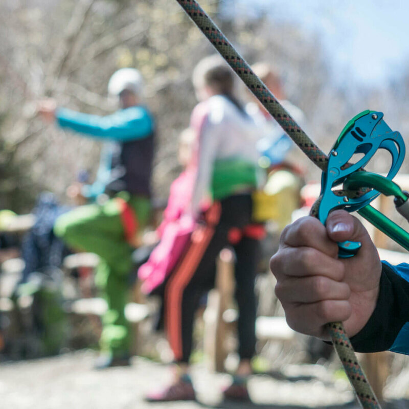 Nahaufnahme von einem Kletter-Sicherungsgerät, einem Seil und bunten Karabinern. Im Hintergrund stehen mehrere Personen.