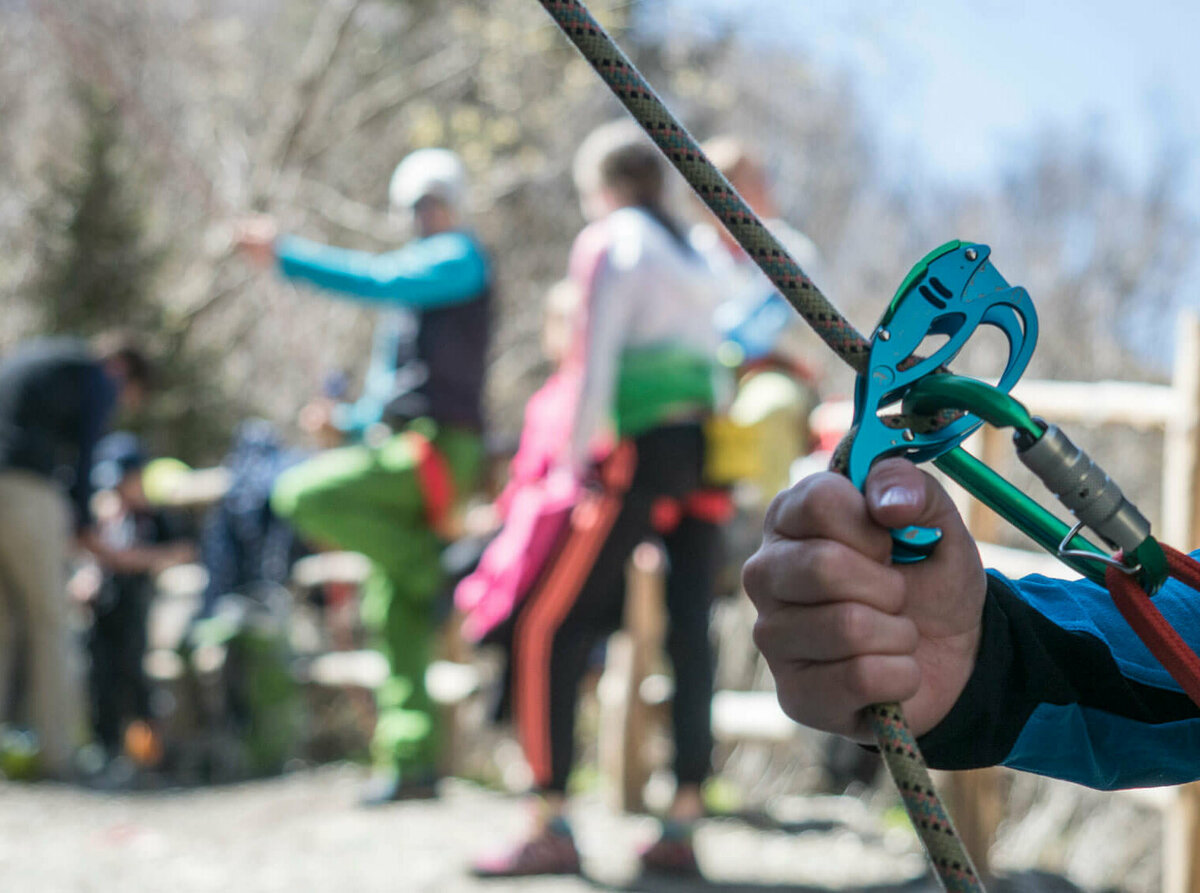 Nahaufnahme von einem Kletter-Sicherungsgerät, einem Seil und bunten Karabinern. Im Hintergrund stehen mehrere Personen.