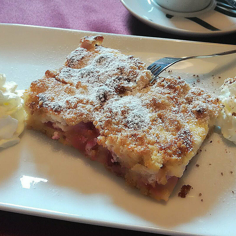 Ein Stück Streuselkuchen auf einem weißen Teller mit zwei Portionen Sahne daneben.