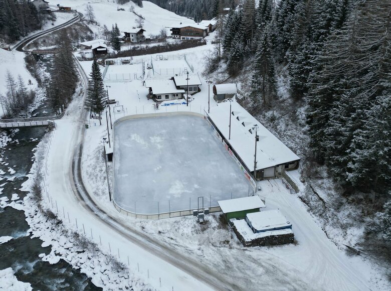 Luftaufnahme vom Eislaufplatz in Prägraten. Links fließt ein Fluss neben der schneebedeckten Straße. Hinter dem Eislaufplatz befindet sich ein Tennisplatz.