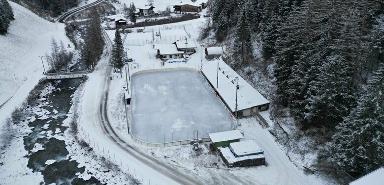 Luftaufnahme vom Eislaufplatz in Prägraten. Links fließt ein Fluss neben der schneebedeckten Straße. Hinter dem Eislaufplatz befindet sich ein Tennisplatz.