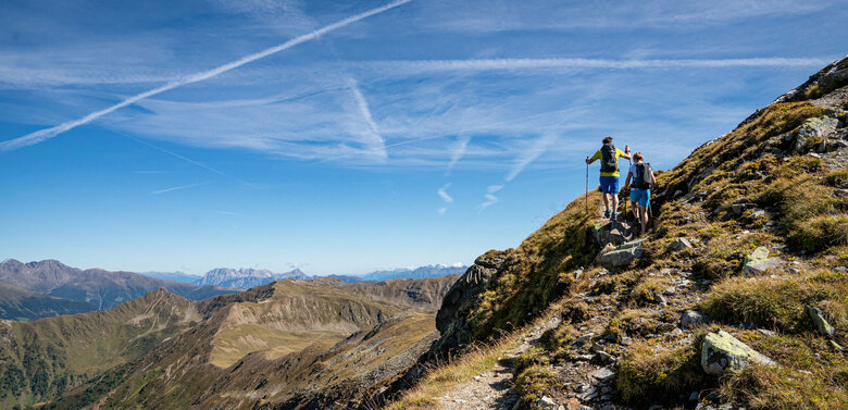 Herz-Ass Villgratental Das Bild zeigt zwei Wanderer auf der Strecke der Herz-Ass-Villgratental Wanderung mit imposanten Blick über die entfernten Berggipfel, die unter strahlend blauen Himmel ruhen.