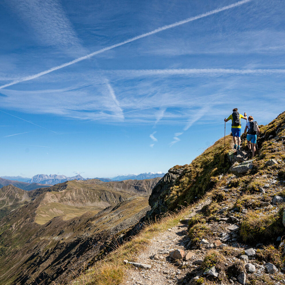 Das Bild zeigt zwei Wanderer auf der Strecke der Herz-Ass-Villgratental Wanderung mit imposanten Blick über die entfernten Berggipfel, die unter strahlend blauen Himmel ruhen.