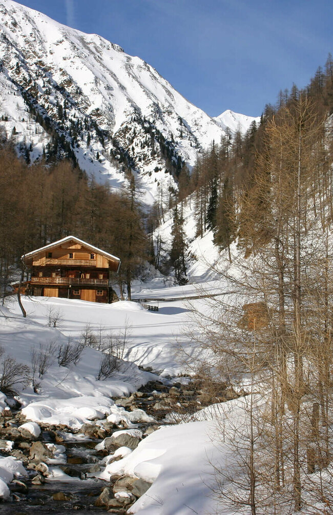 Eine urige, hölzerne Almhütte liegt in der verschneiten Landschaft des Winkeltals in Außervillgraten. Im Hintergrund heben sich die bewaldeten und schneebedeckten Bergwände empor.