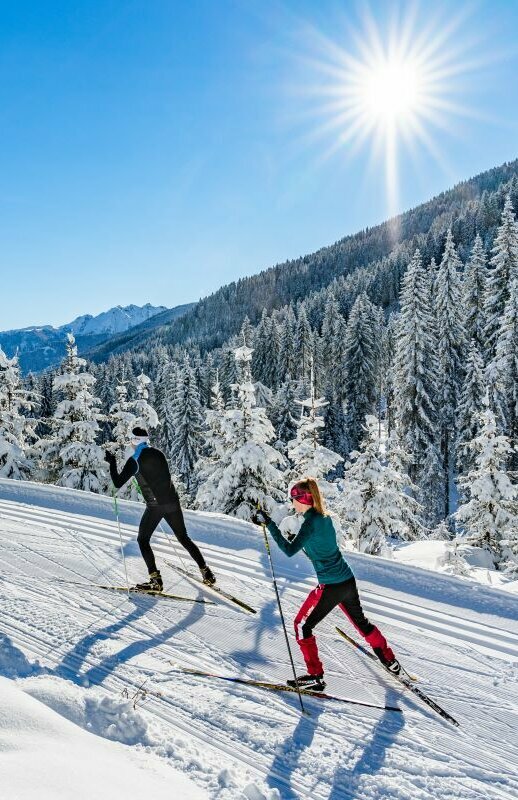 Zwei Langläufer:innen laufen bei strahlendem Sonnenschein auf der Loipe in Obertilliach mit einem frisch verschneiten Wald im Hintergrund