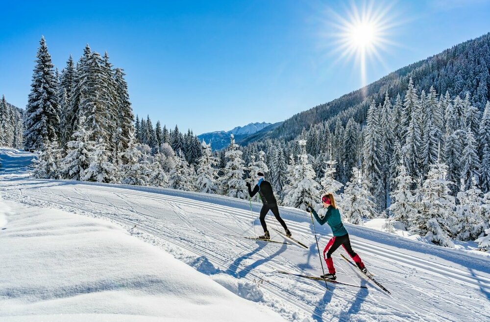 Zwei Langläufer:innen laufen bei strahlendem Sonnenschein auf der Loipe in Obertilliach mit einem frisch verschneiten Wald im Hintergrund