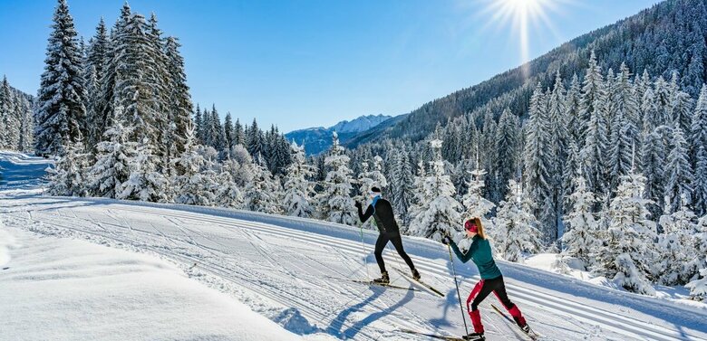 Zwei Langläufer:innen laufen bei strahlendem Sonnenschein auf der Loipe in Obertilliach mit einem frisch verschneiten Wald im Hintergrund