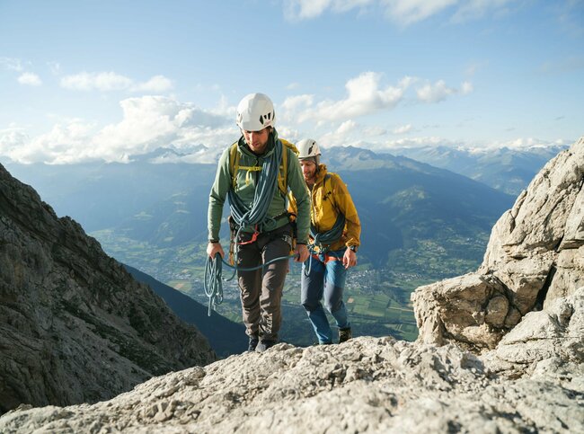 Zwei Kletterer mit weißen Helmen und blauen Seilen bei der Besteigung Laserz, mit Blick auf das Tal dahinter.