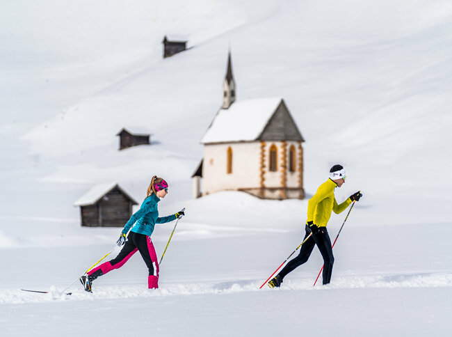 Zwei Langläufer:innen laufen in den Tilliacher Feldern mit einigen kleinen Heustadeln und der Filialkirche St. Nikolaus im Hintergrund
