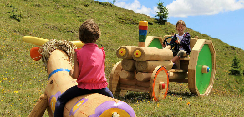 Zwei Mädchen, die auf einer bunten Holzkuh bzw. auf einem bunten Holztraktor sitzen. Dahinter ein grasbewachsener Berghang mit spärlichem Bewuchs und einem leicht bewölkten Himmel.