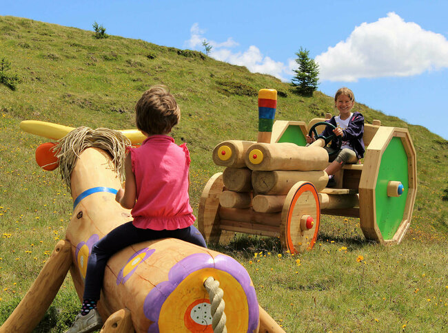 Zwei Mädchen, die auf einer bunten Holzkuh bzw. auf einem bunten Holztraktor sitzen. Dahinter ein grasbewachsener Berghang mit spärlichem Bewuchs und einem leicht bewölkten Himmel.